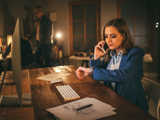 junge Frau mit Smartphone am Ohr sitzt spät Abends im Büro und schaut auf ihre Uhr